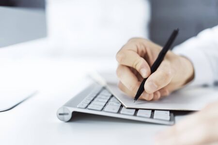 Closeup of man hand writing in diary on a table with computer. Focused on a pen. Close upの写真素材