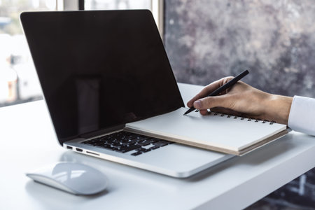 Man writes with a pen in notebook on laptop keyboard in a sunny office, business and education...