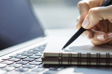 Woman writes with a pen in notebook on laptop keyboard in a sunny office, business and education...