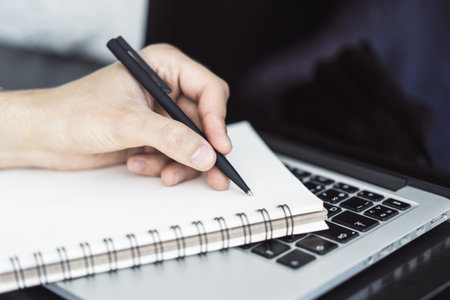 Man writes with a pen in diary on laptop keyboard in a sunny office, business and education concept....
