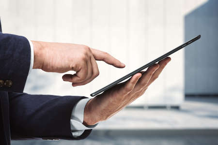 Businessman holds digital tablet and clicks on screen near the exterior of the business center on a sunny day, close up. Search conceptの写真素材