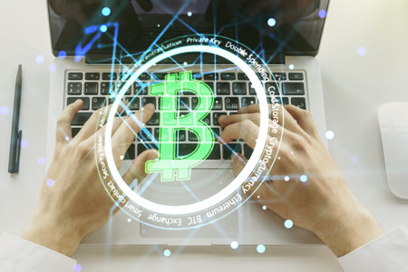 Double exposure of creative Bitcoin symbol hologram with hands typing on computer keyboard on...