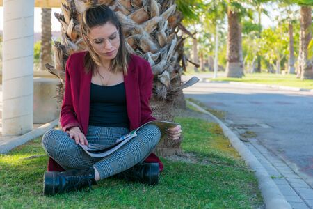 Young woman with glasses sitting on a park grass next to a palm tree reading a magazineの写真素材