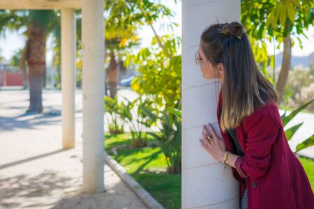 Young woman with glasses and standing out behind a column in a parkの写真素材