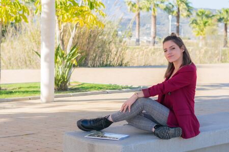 Attractive girl with red jacket sitting on a bench in the park.の写真素材