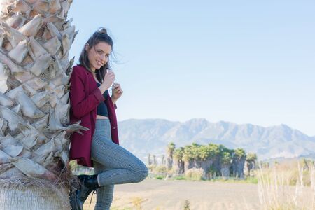 Young and friendly woman posing next to a palm tree with mountains and clear sky in the backgroundの写真素材