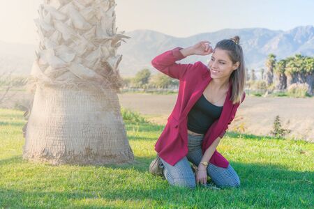 Cheerful young woman kneeling on the grass next to a palm tree with her hand on her forehead looking to the sideの写真素材