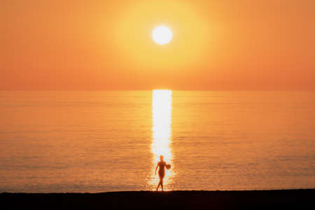 Young boy with a ball in his hand on the beach passing by the reflections of the sun on a sunset.の写真素材