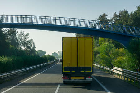 Trailer with a closed box and yellow color driving on a highway and passing under a pedestrian bridge.の写真素材
