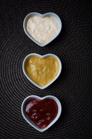 Small heart-shaped bowls with ketchup, mayonnaise and mustard sauce, vertical shot.