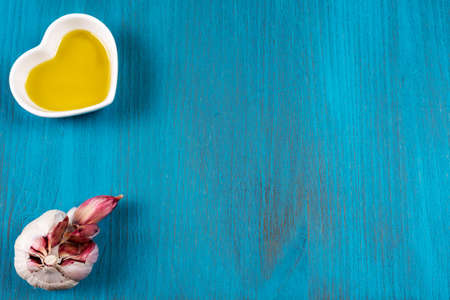 Blue wooden background with a small heart-shaped bowl with olive oil and a head of garlic.の写真素材