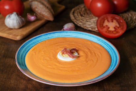 Round plate with salmorejo, ham and egg, along with some tomatoes, bread and garlic, ingredients used for its preparation.の写真素材