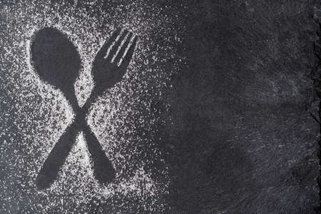 Crossed spoon and fork figure created in sugar relief on a black stone background.の写真素材