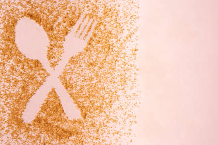 Crossed spoon and fork figure created in relief of muscovado sugar on pastel pink background.