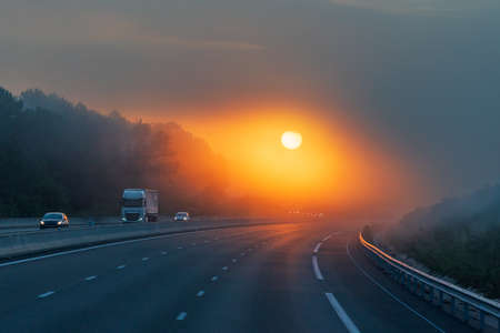 Landscape of a highway with a truck and several vehicles circulating at dawn, and the sun appearing through the clouds, allowing its circumference to be seen through the mist.の写真素材
