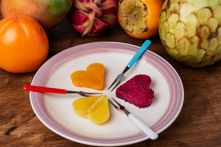 Rectangular plate with three pieces of tropical fruits in the shape of a heart such as pitahaya, mango and a persimmon, next to the plate the pieces of whole fruits.の写真素材