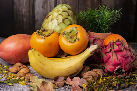Still life of tropical fruits such as mango, persimmon, custard apple, banana and pitahaya surrounded by almonds, walnuts and dry leaves.の写真素材