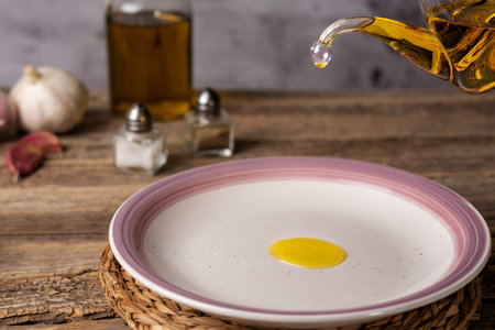 Extra virgin olive oil pouring from an oil can into a round white plate, on a rustic wooden table.の写真素材