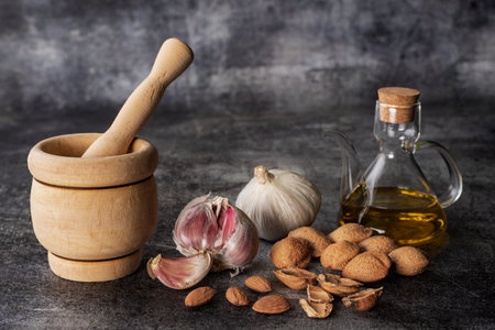 Wooden mortar, kitchen utensil to mash, next to some garlic, an oil can and almonds, on a gray stone background.の写真素材