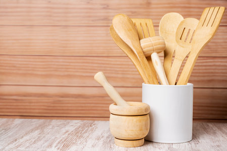 Wooden kitchen utensils inside a jar and next to a mortar, spoon, fork, colander and tongs among others, arranged in a decorative way with a light wooden background.の写真素材
