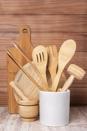 Wooden kitchen utensils, plate, cutting board and mortar, next to a jar with a spoon, fork and tongs among others, decoratively arranged with a light wooden background.の写真素材