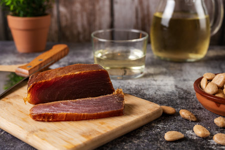 Piece of tuna mojama or cecina cut into pieces on a cutting board accompanied by almonds, and a glass and a jug with white wine.の写真素材