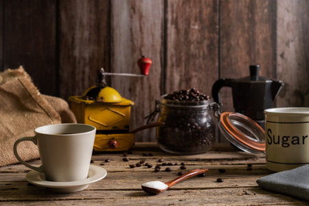 Cup of fresh coffee next to a wooden spoon with sugar, in the background an old coffee grinder, an Italian coffee maker and a jar with coffee.の写真素材