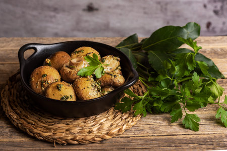 Plate of baked mushrooms with garlic, parsley and Provencal herbs dressing, on a rustic table with plant leaves for cooking.の写真素材