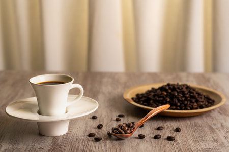 Cup of coffee next to a plate and a wooden spoon with roasted coffee beans, on a light wooden table.の写真素材