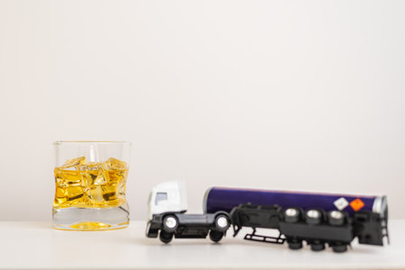 Toy tanker truck with danger labels for dangerous goods overturned next to a glass of whiskey, alcohol incompatibility and professional driving.の写真素材