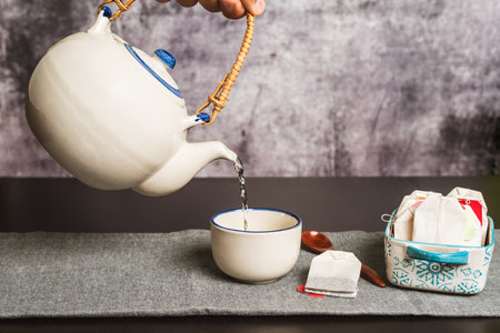 A hand holding a teapot while pouring hot water to prepare the tea, along with several sachets with...
