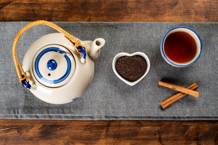 Porcelain teapot next to a small heart-shaped bowl with ground tea, a cup with tea and cinnamon sticks.の写真素材