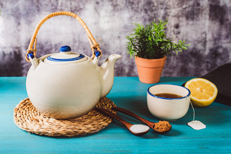 Porcelain teapot next to a cup of tea and half a lemon, with two wooden spoons, one with white sugar and the other with brown sugar.の写真素材