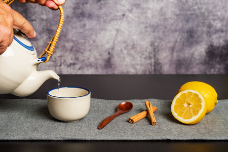 Some hands holding a teapot while pouring the hot water to prepare the tea, along with some pieces of cinnamon and lemons.の写真素材