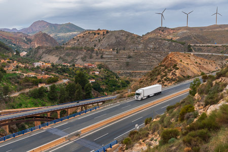 Truck with a refrigerated semi-trailer driving on a highway and a landscape with bridges, mountains, towns and wind turbines.の写真素材