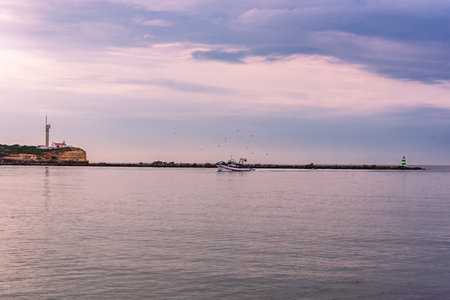 Lighthouse at Punta del Altar, Ferragudo, with a fishing boat entering the ria. Algarve.の写真素材