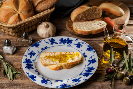 Slice of bread with olive oil on a plate next to a cutting board with more slices, an oil can, garlic, branches with olives, salt, pepper and a bread basket with bread.の写真素材
