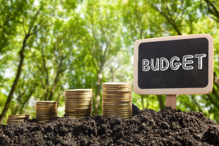 Time is money financial opportunity concept. Golden coins in soil with Chalkboard with Budget word on blurred urban backgroundの写真素材