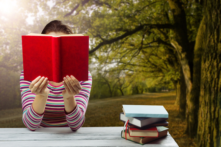 Young woman reading a book and covering her face ,sitting by wooden table with stack of colorful hardback books on blurred nature landscape backdropの写真素材