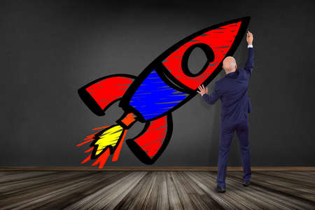 View of a Businessman in front of a wall drawing a colorfull start up rocket - business conceptの写真素材
