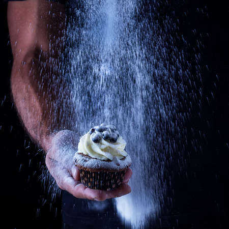 Cupcake in mans hand and the pouring icing sugar.Selective focus,dark background.の写真素材