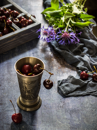 Metal glass with cherry,box with sweet cherry and blue flowers on dark blue background. Vintage style. Selective focus.の写真素材