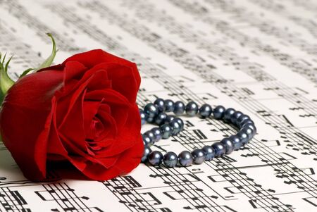 A single red rose lays on top of a complicated score of music. A gift of black pearls sits next to the rose.の写真素材