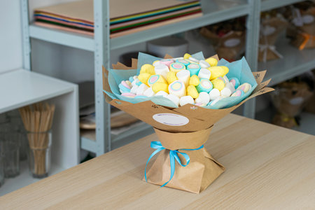 Creative bouquet made of colorful marshmallows wrapped in craft paper with a blue ribbon, displayed on a wooden table in a cozy workshop or gift shop. Bright and cheerful composition ideal for celebrations or gift ideasの写真素材