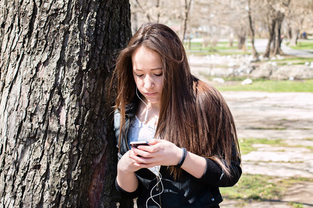 Young girl with smart phone leaning on the old big tree in the parkの写真素材