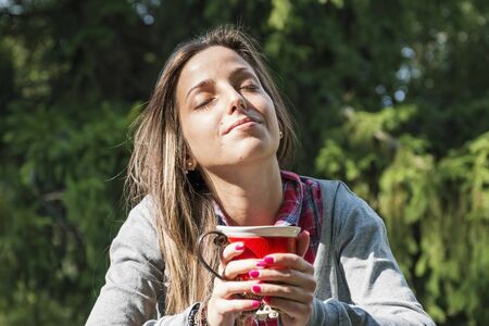 Beautiful young woman with a red cup drinking coffee in a morning parkの写真素材