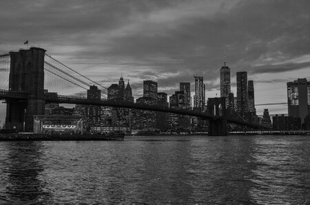 Black and white photo of the Brooklyn Bridge during a cloudy sunset in foreground with New York City skyline in background.の写真素材