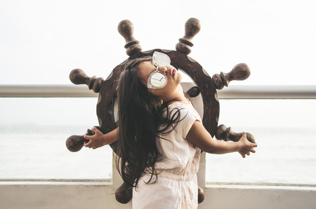 Little girl playing to be a sailor on a balcony with a boat rudder and the background of the seaの写真素材