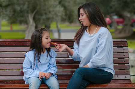 Mother and her little daughter sitting on a bench eating a cookie in the parkの写真素材