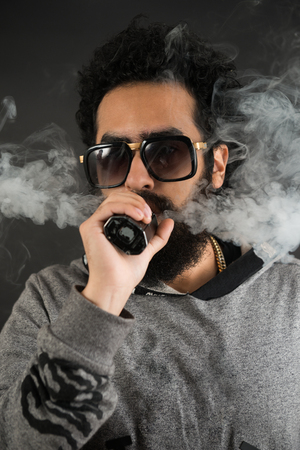 Portrait of a young guy with big beard and sunglasses and vaping an electronic cigarette on a black background.の写真素材